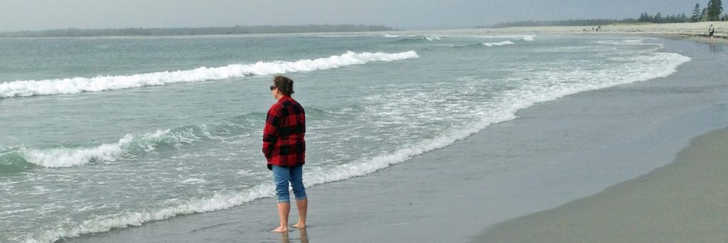 Deirdre at a beach in Nova Scotia. She is standing on sand looking out at the waves rolling in